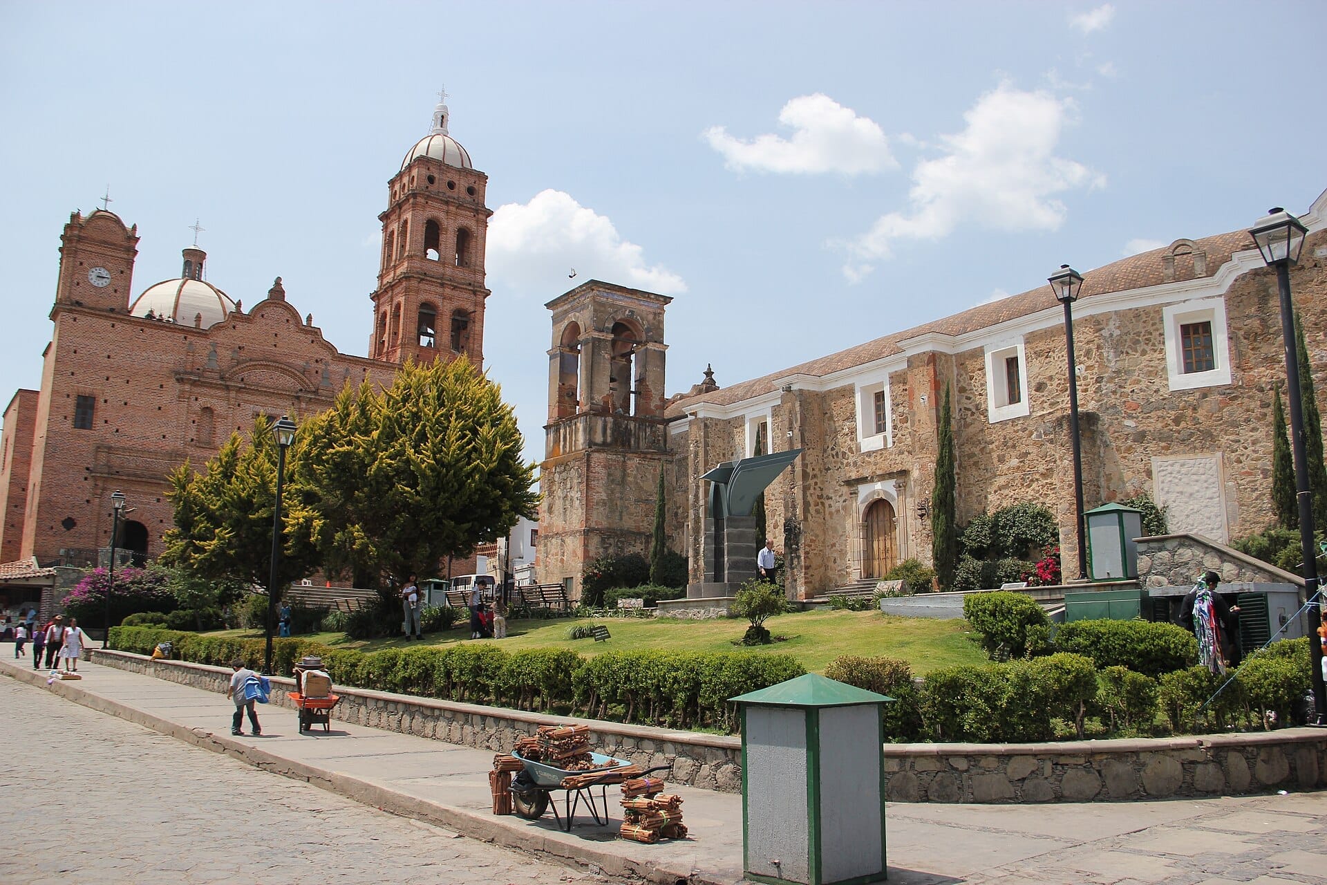 Piazza storica di Tapalpa con chiesa coloniale in mattoni, edifici in pietra, giardini verdi e persone che godono di una giornata di sole a Jalisco.