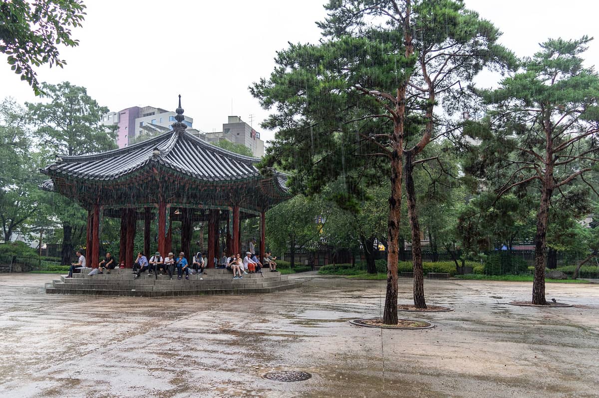 O Parque Tapgol em Seul num dia de chuva, mostrando o pavilhão histórico com pessoas sentadas embaixo, árvores ao redor e prédios da cidade ao fundo.