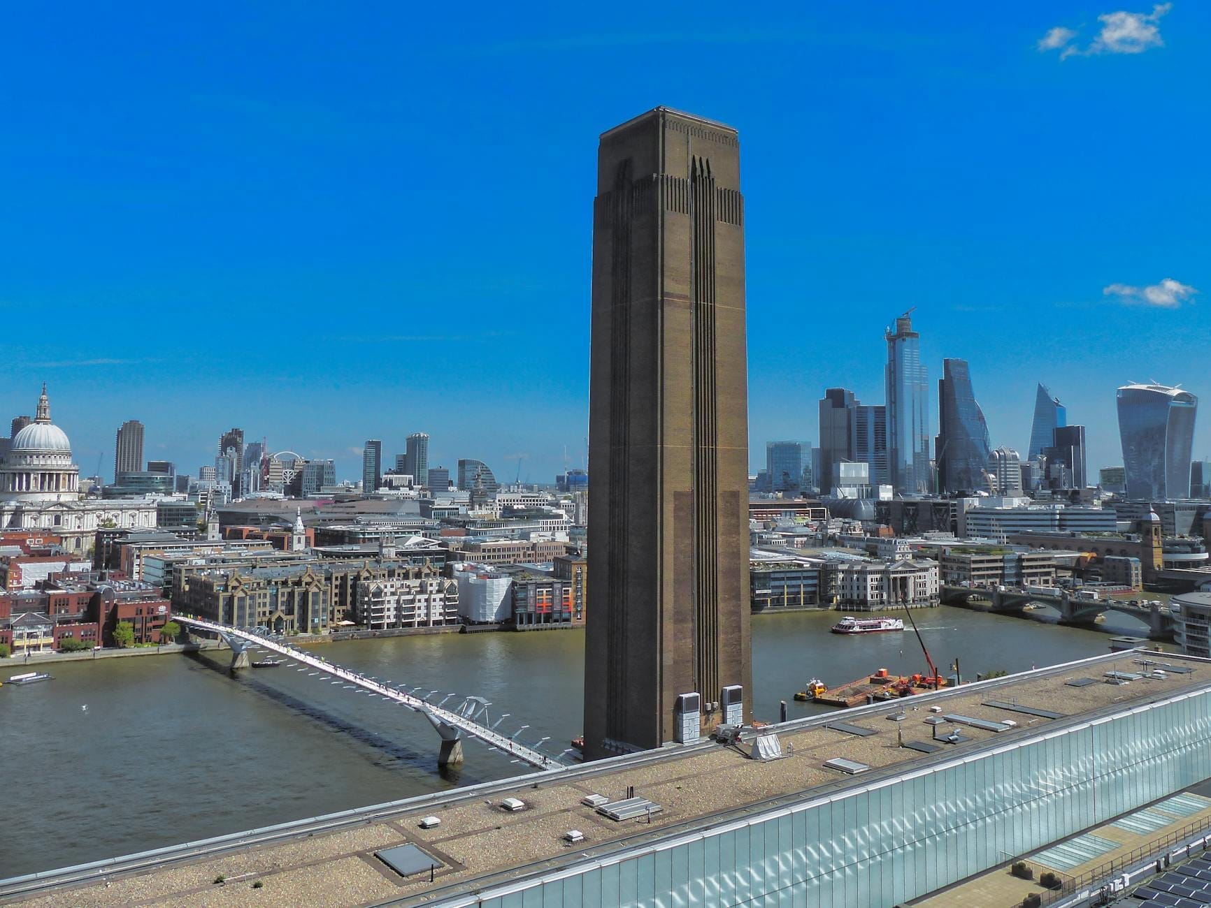 Vista della caratteristica ciminiera della Tate Modern con il Tamigi, il Millennium Bridge e lo skyline di Londra sotto un cielo azzurro brillante.