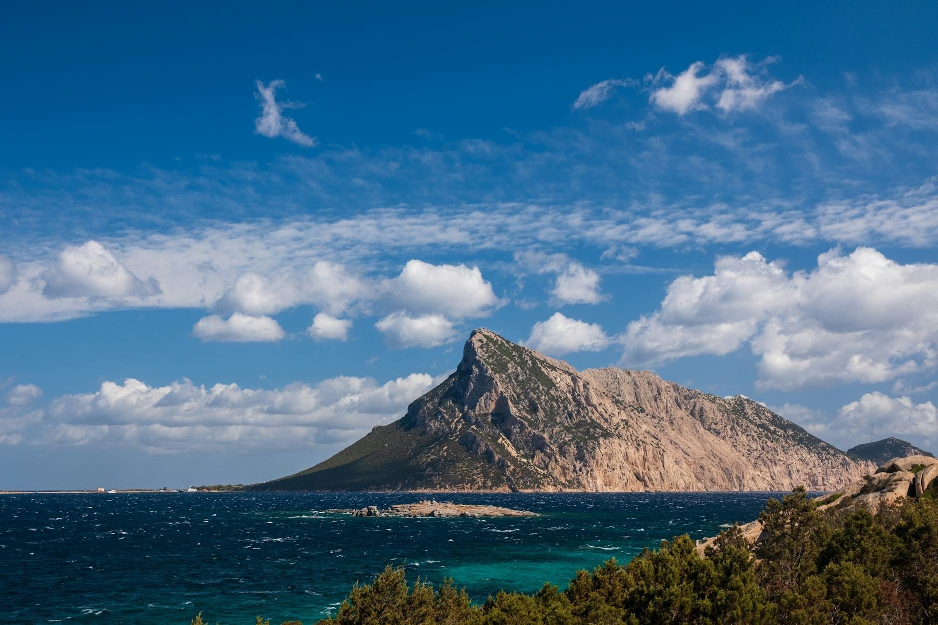 Die schroffe Kalksteininsel Isola Tavolara erhebt sich steil aus dem tiefblauen Tyrrhenischen Meer, mit vereinzelten Wolken und wilder Küstenlandschaft im Vordergrund.