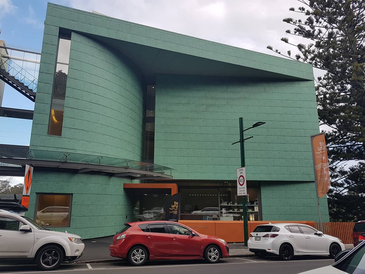 Außenansicht der Te Uru Waitākere Contemporary Gallery mit ihren markanten grün geschwungenen Wänden und dem orangefarbenen Eingang, aufgenommen von der Straße in Titirangi, Auckland.
