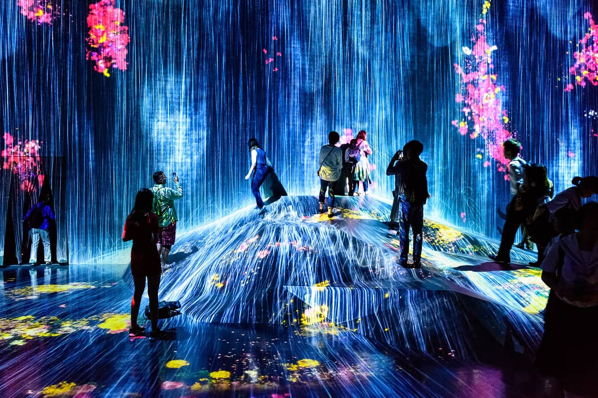 Visitors explore teamLab Borderless Tokyo, surrounded by immersive digital waterfall and floral projections cascading over walls and floors in vivid colors.
