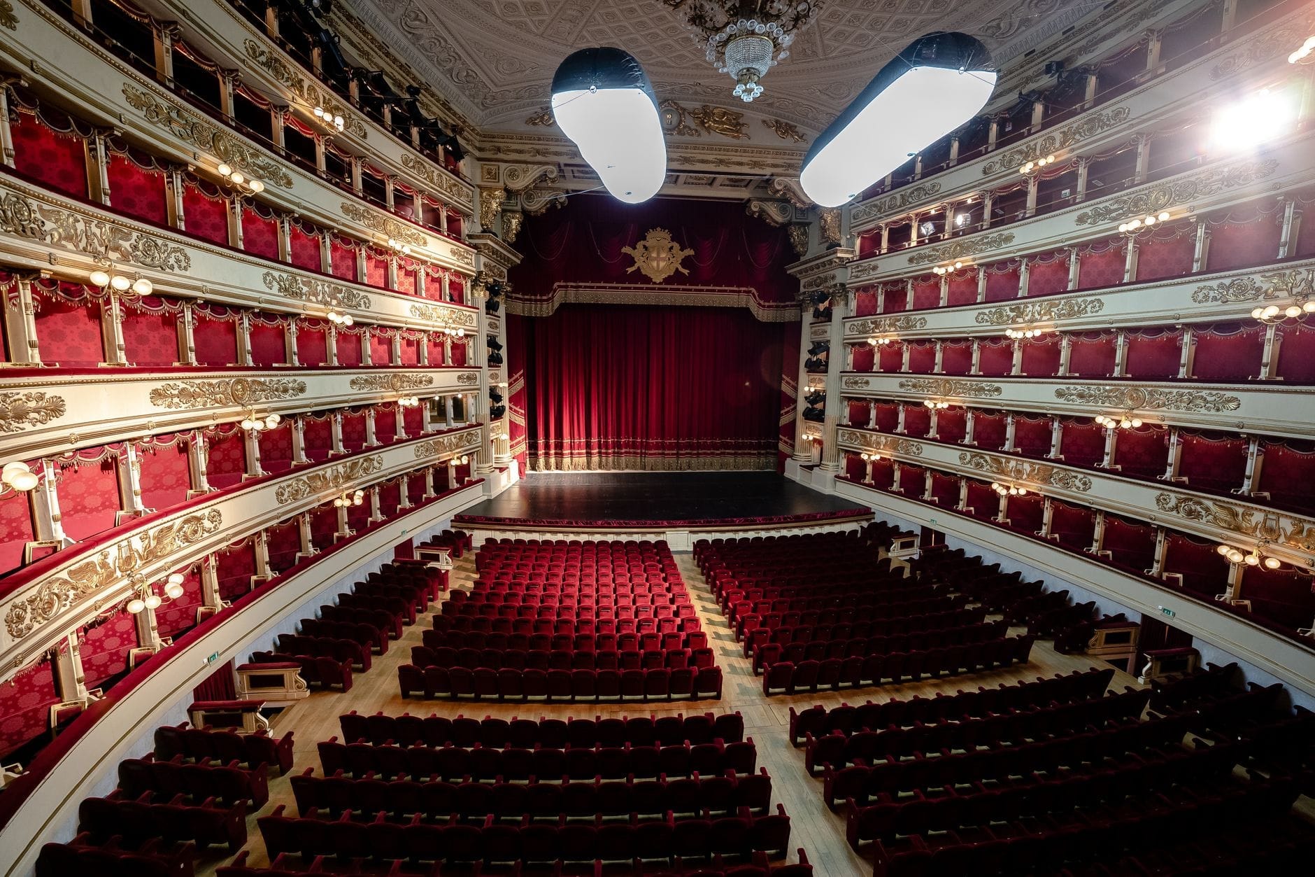 Vista interna del Teatro alla Scala con le poltroncine in velluto rosso, i palchi dorati disposti a ferro di cavallo e il grande sipario rosso calato sul palcoscenico sotto un'illuminazione elegante.