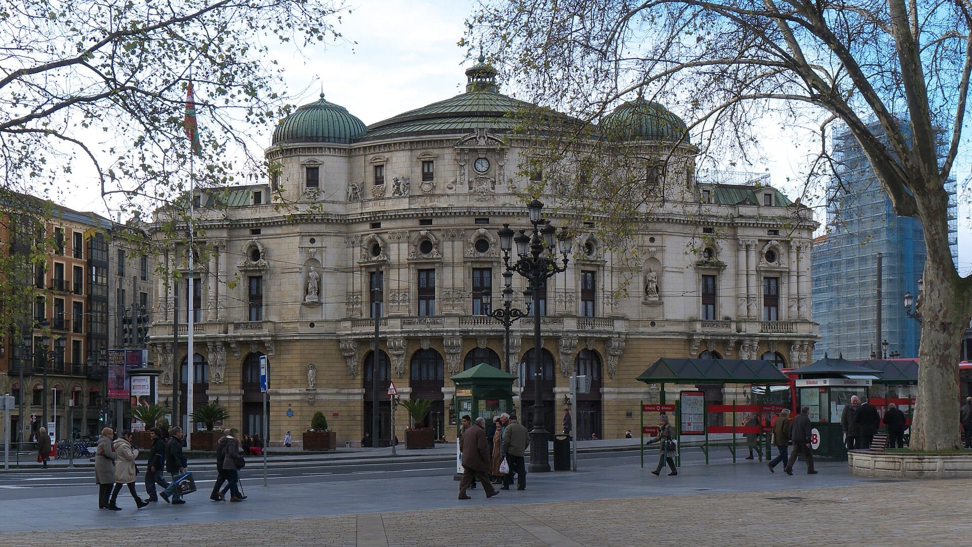 Front view of Teatro Arriaga in Bilbao with its ornate neo-baroque facade, people walking by, and trees framing the scene.