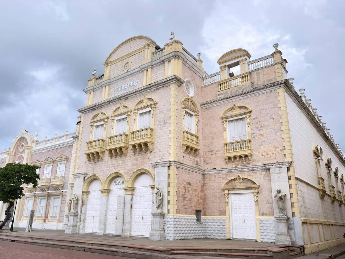 Teatro Heredia in Cartagena features ornate pastel facades, arched doors, and statues under a cloudy sky, showcasing early-20th-century architecture.