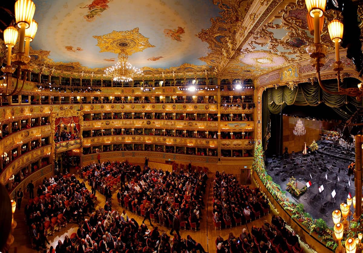 Weitwinkelansicht des prachtvoll vergoldeten Zuschauerraums im Teatro La Fenice, gefüllt mit Publikum vor einer Bühne, die für eine Vorstellung eingerichtet ist, darunter eine aufwendig bemalte Decke.