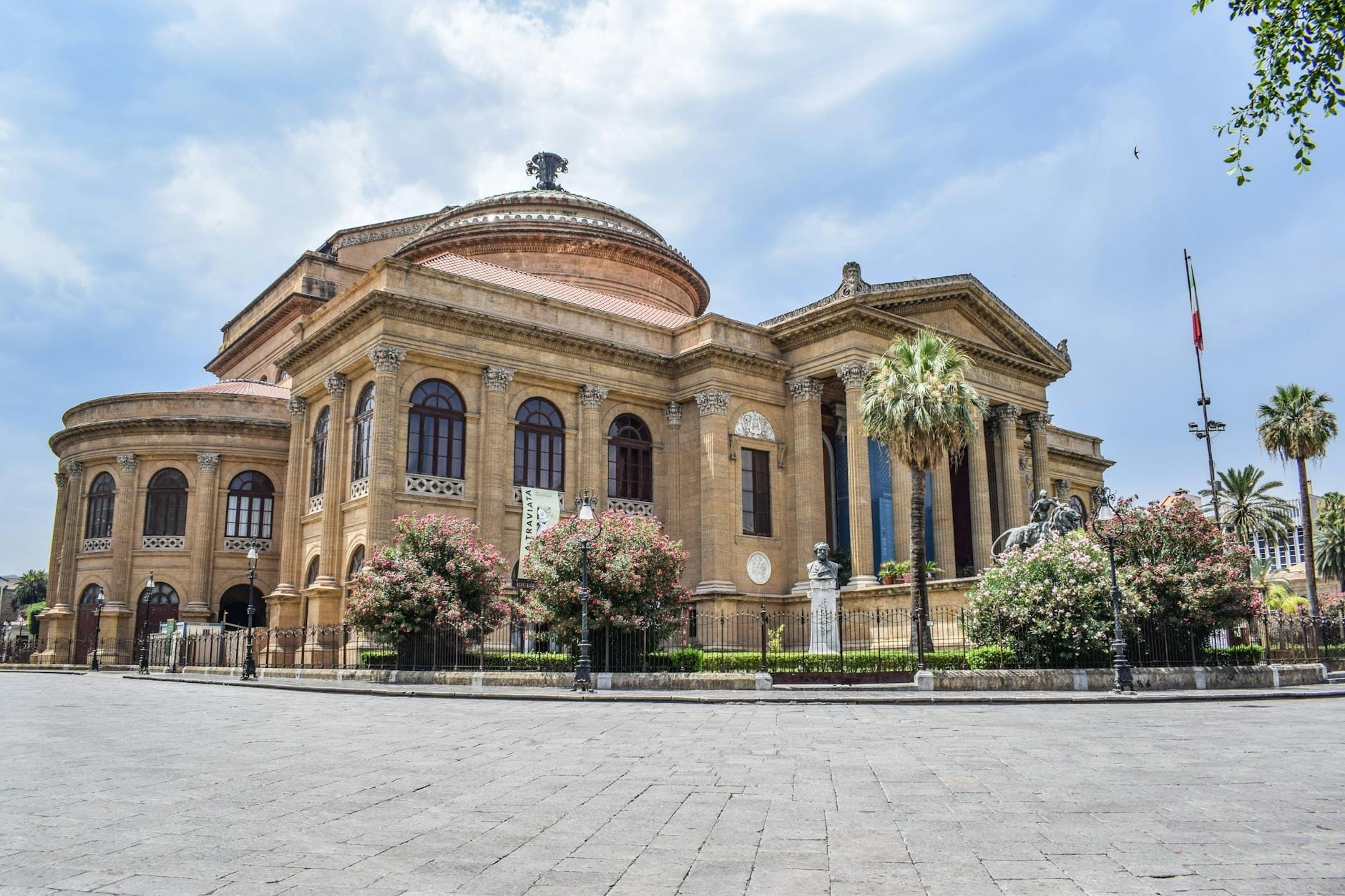 Weitwinkelperspektive auf das neoklassische Teatro Massimo in Palermo, eingerahmt von Palmen und buntem Blattwerk unter strahlend blauem Himmel.