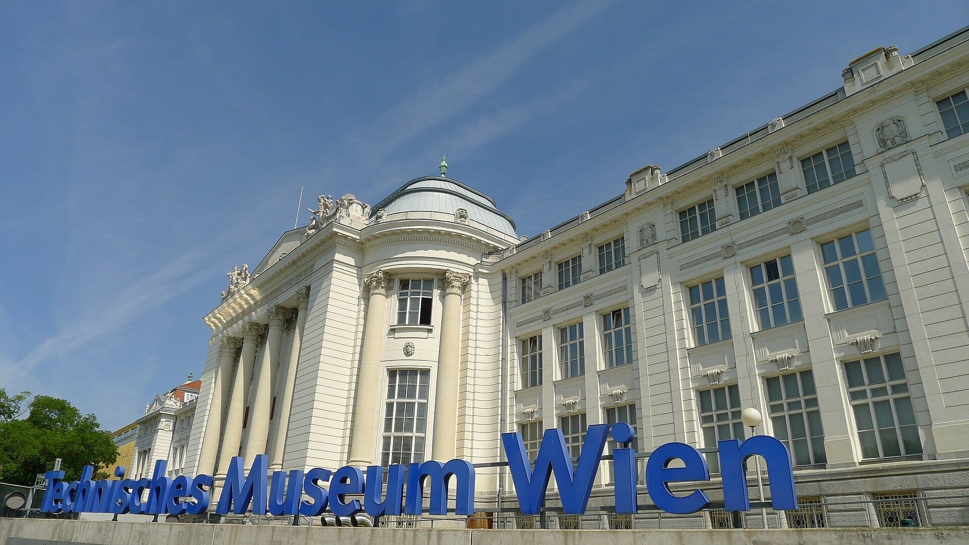Die prachtvolle weiße Fassade des Technisches Museum Wien mit hohen Säulen, Statuen und großer blauer Beschilderung unter einem klaren, sonnigen Himmel.