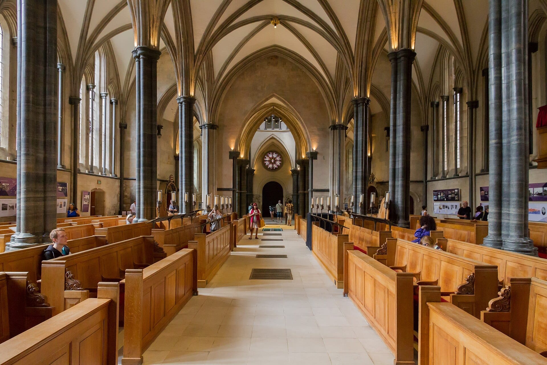 Vista interna della Temple Church a Londra, con imponenti archi gotici, colonne in pietra, banchi in legno, vetrate colorate e diversi visitatori.
