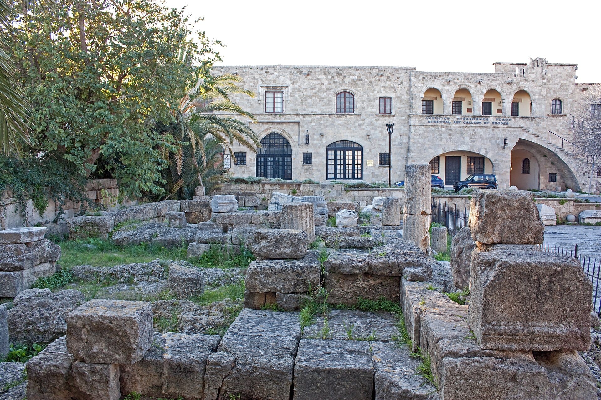 ซากหินของวิหาร Aphrodite ใน Rhodes Old Town พร้อมเสาหินที่ผุกร่อนและบล็อกโบราณที่ล้อมรอบด้วยพืชพรรณและอาคารประวัติศาสตร์