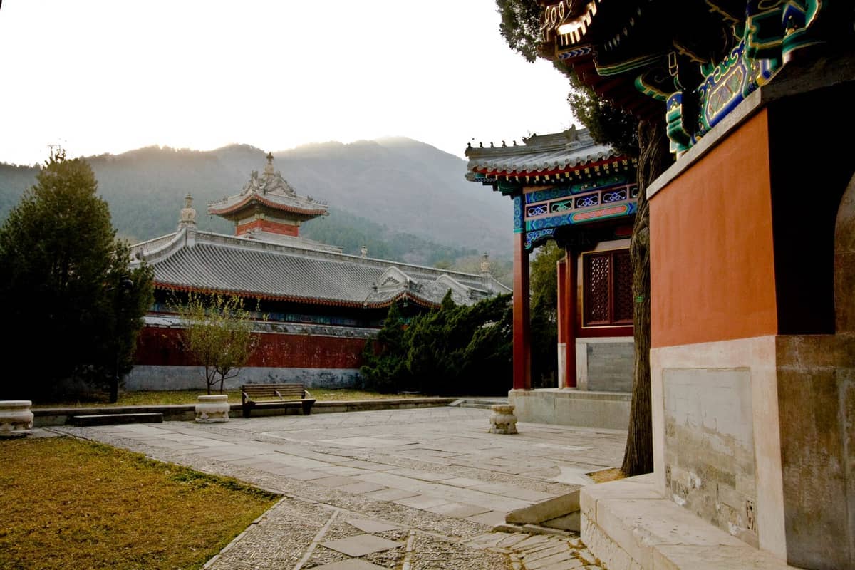 Innenhof des Tempels der Azurblauen Wolken mit verzierten rot-grauen Gebäuden, dekorierten Dachlinien, Kiefern und dunstiger Bergkulisse in Peking.