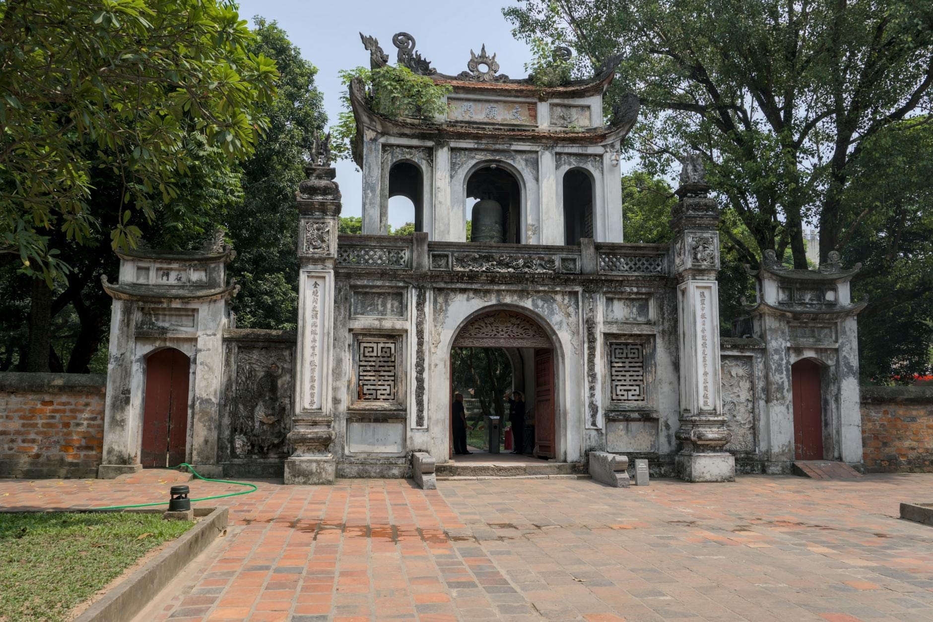 Wide view of the main gate of the Temple of Literature in Hanoi with traditional Vietnamese architecture, set among trees and a brick courtyard under clear daylight.