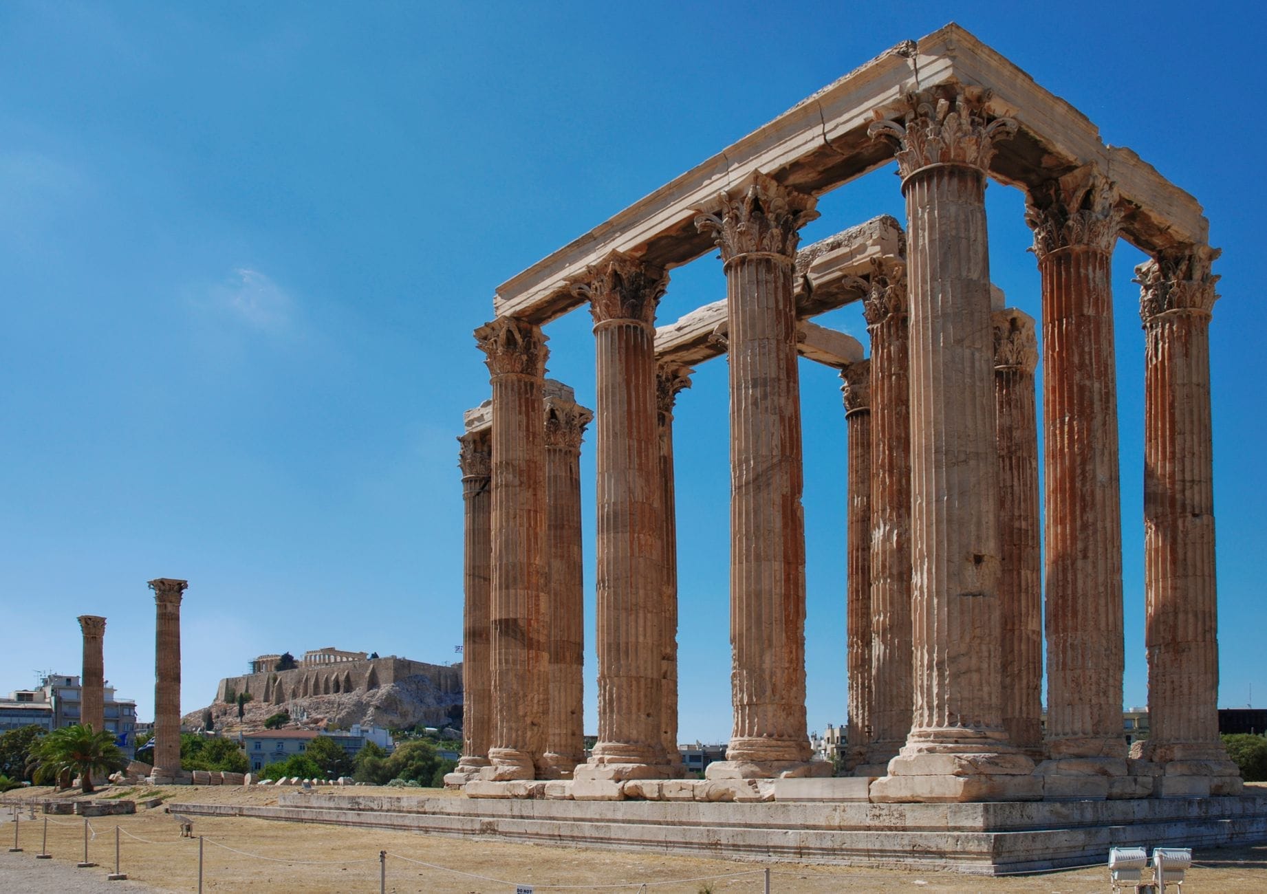 Le imponenti colonne corinzie del Tempio di Zeus Olimpio si innalzano sotto un cielo azzurro ad Atene, con l'Acropoli visibile sullo sfondo.