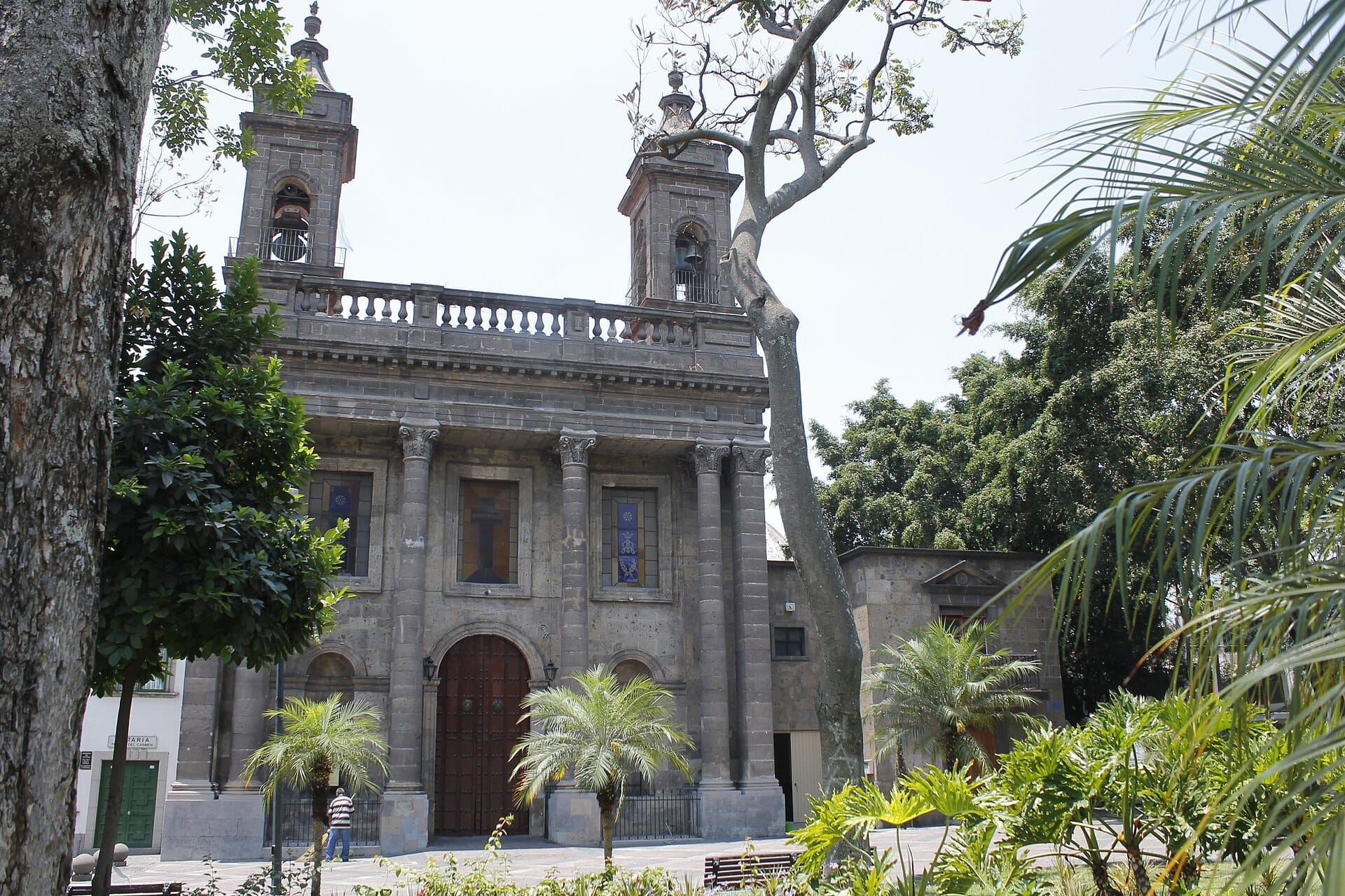Vista frontale del Templo del Carmen, una chiesa in pietra neoclassica incorniciata da palme e vegetazione nel centro storico di Guadalajara.
