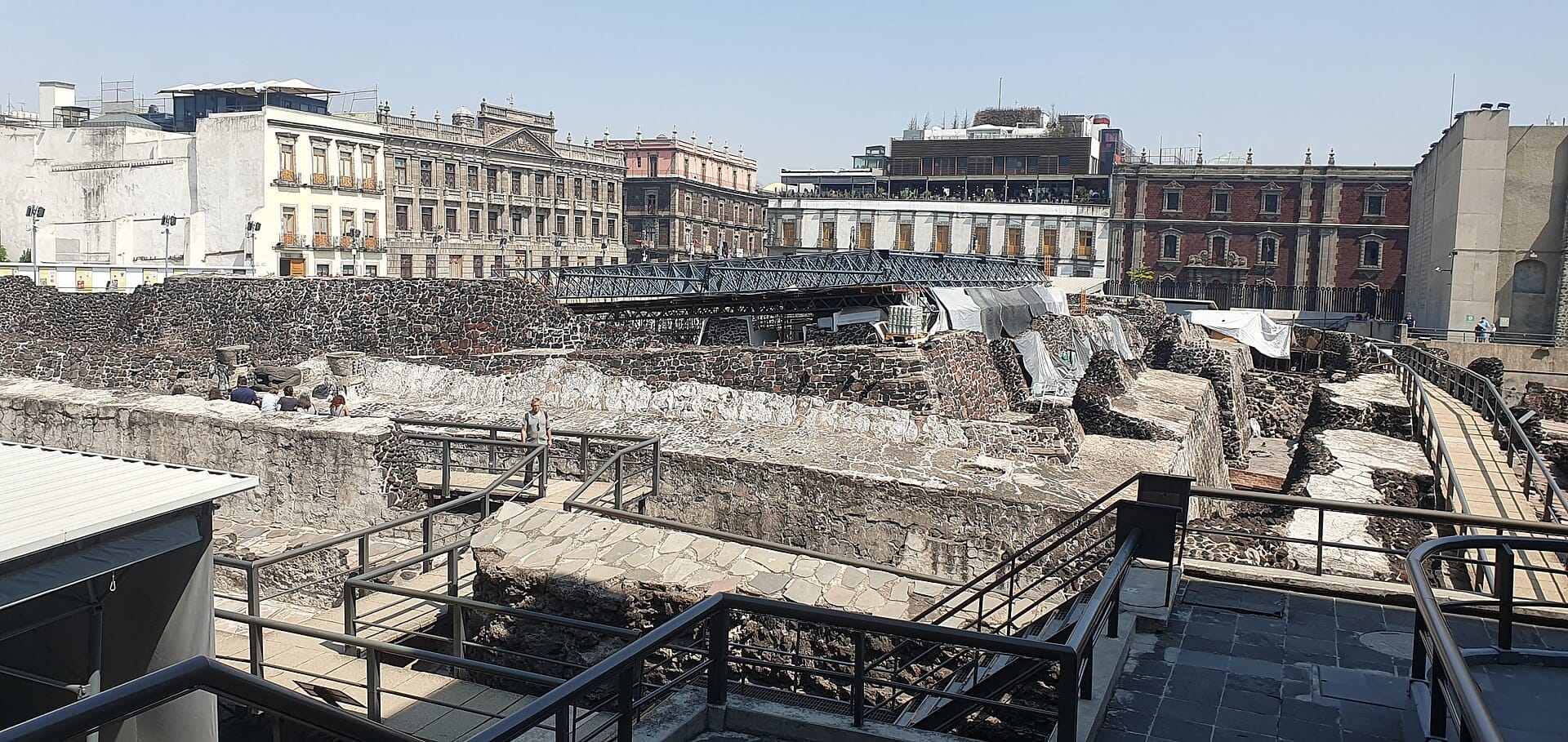 Rovine in pietra del Templo Mayor con passerelle e visitatori, circondate da edifici storici sotto un cielo azzurro limpido a Città del Messico.