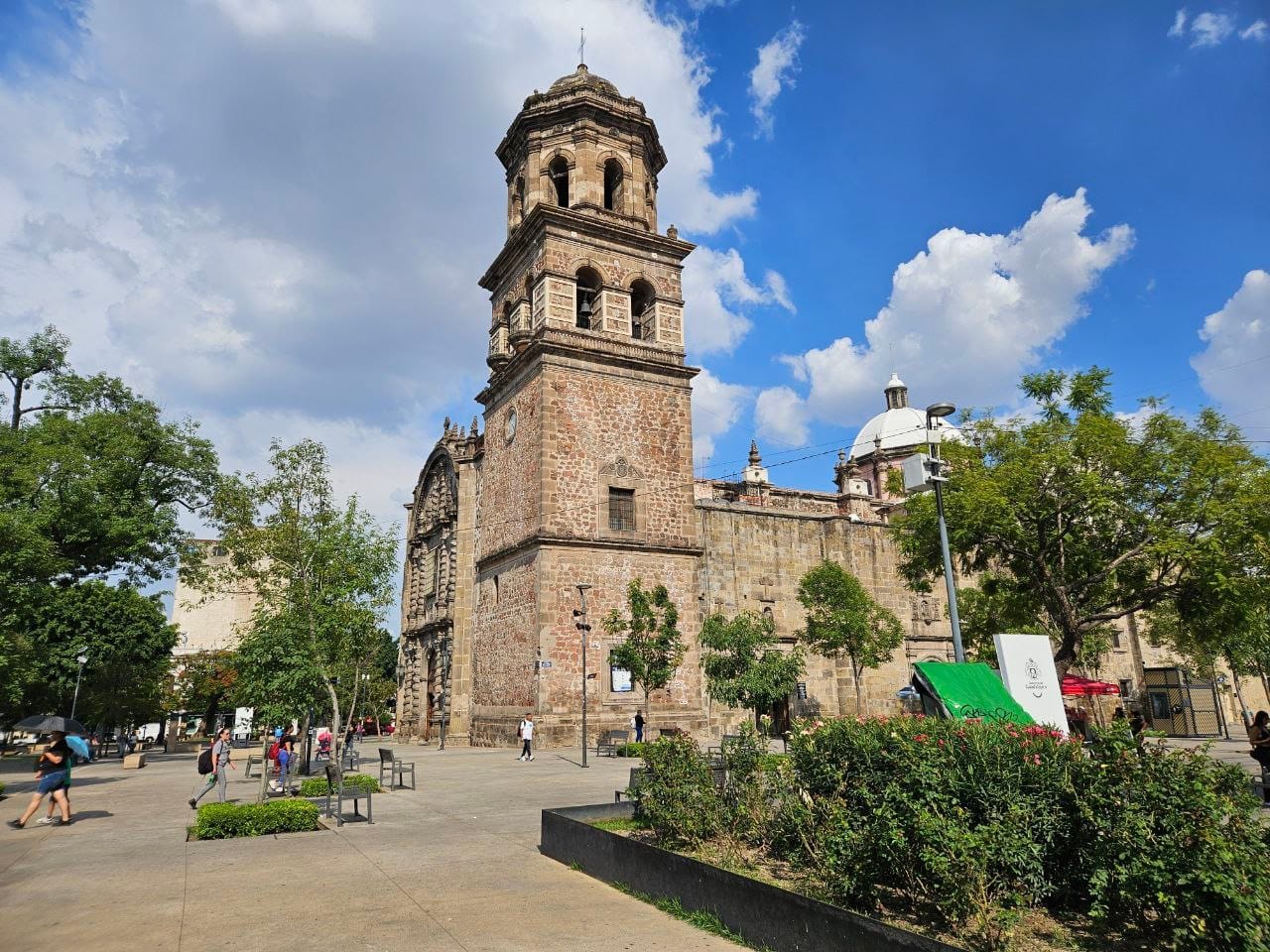Il Templo de San Francisco de Asís a Guadalajara svetta con la sua storica torre in pietra, circondato da vegetazione e visitatori in una giornata di sole.