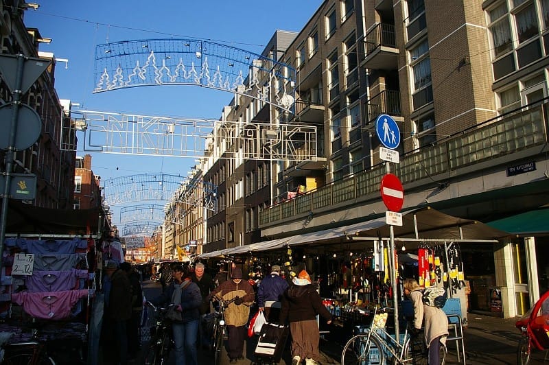 Compradores exploran puestos de mercado al aire libre bajo el letrero de Ten Katemarkt en una calle soleada bordeada de modernos edificios de apartamentos en Ámsterdam.
