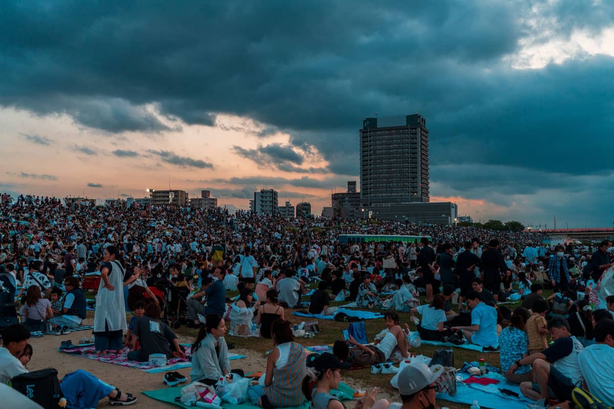 Large crowds sitting on picnic mats along a riverside embankment in a city at dusk, waiting for a festival or fireworks event.