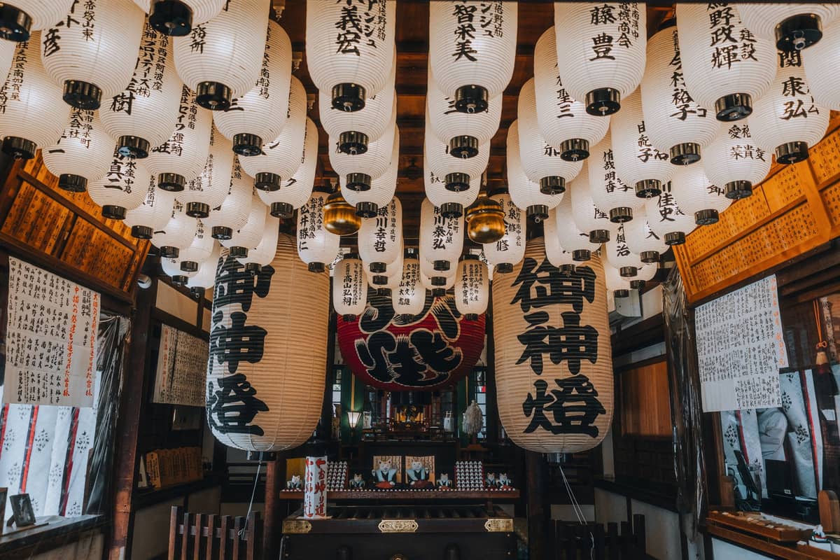 Rows of Japanese lanterns with calligraphy hang inside a shrine, with ritual offerings and a central altar visible beneath.