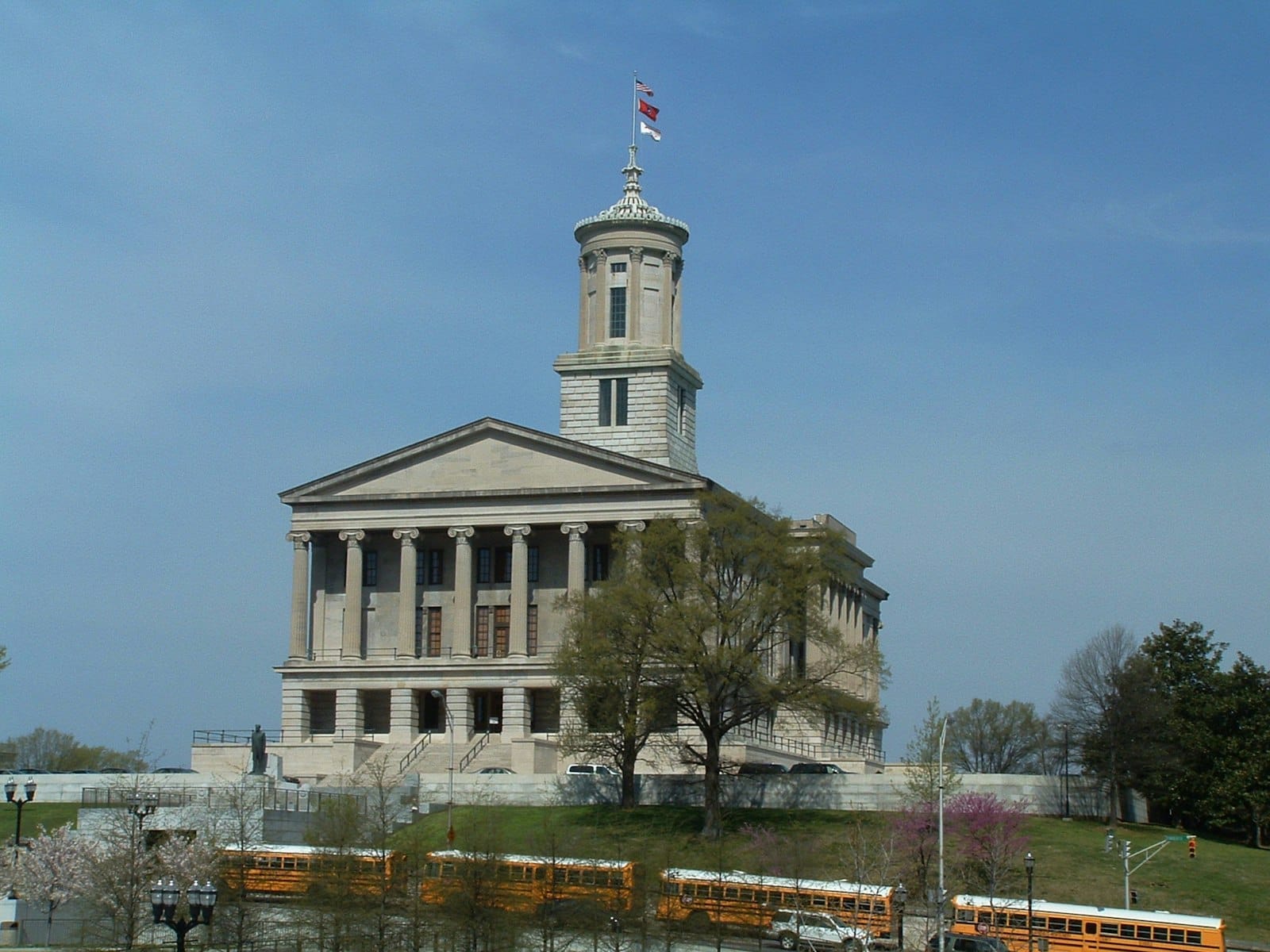 Vista panorámica del Capitolio del Estado de Tennessee bajo un cielo azul, con árboles verdes y autobuses escolares amarillos en primer plano.