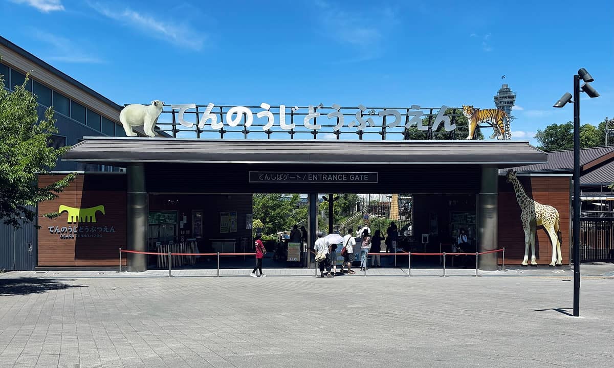 Wide view of the main entrance gate to Osaka Tennoji Zoo under a blue sky, with animal statues and families entering.
