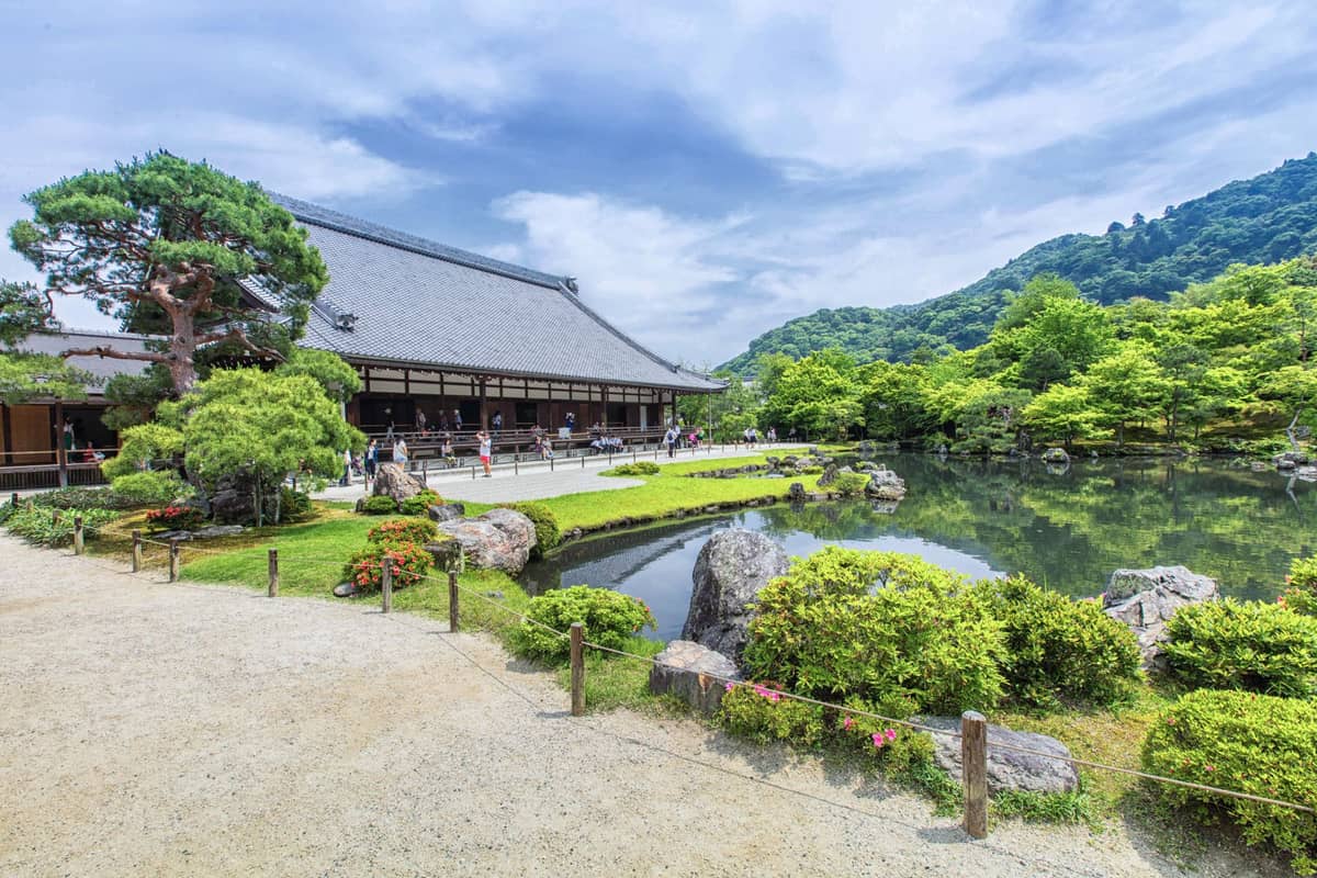 Wide view of Tenryu-ji Temple’s main hall and iconic Sogenchi Zen garden pond, with manicured greenery and Arashiyama mountains in the background on a sunny day.