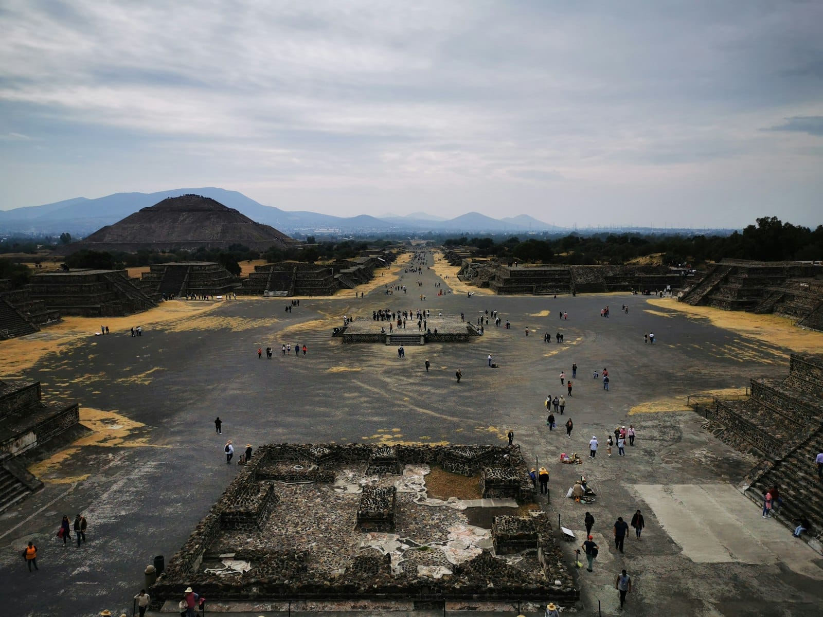 Vista ao longo da Avenida dos Mortos em Teotihuacán, com visitantes caminhando entre ruínas antigas e a Pirâmide da Lua ao fundo.