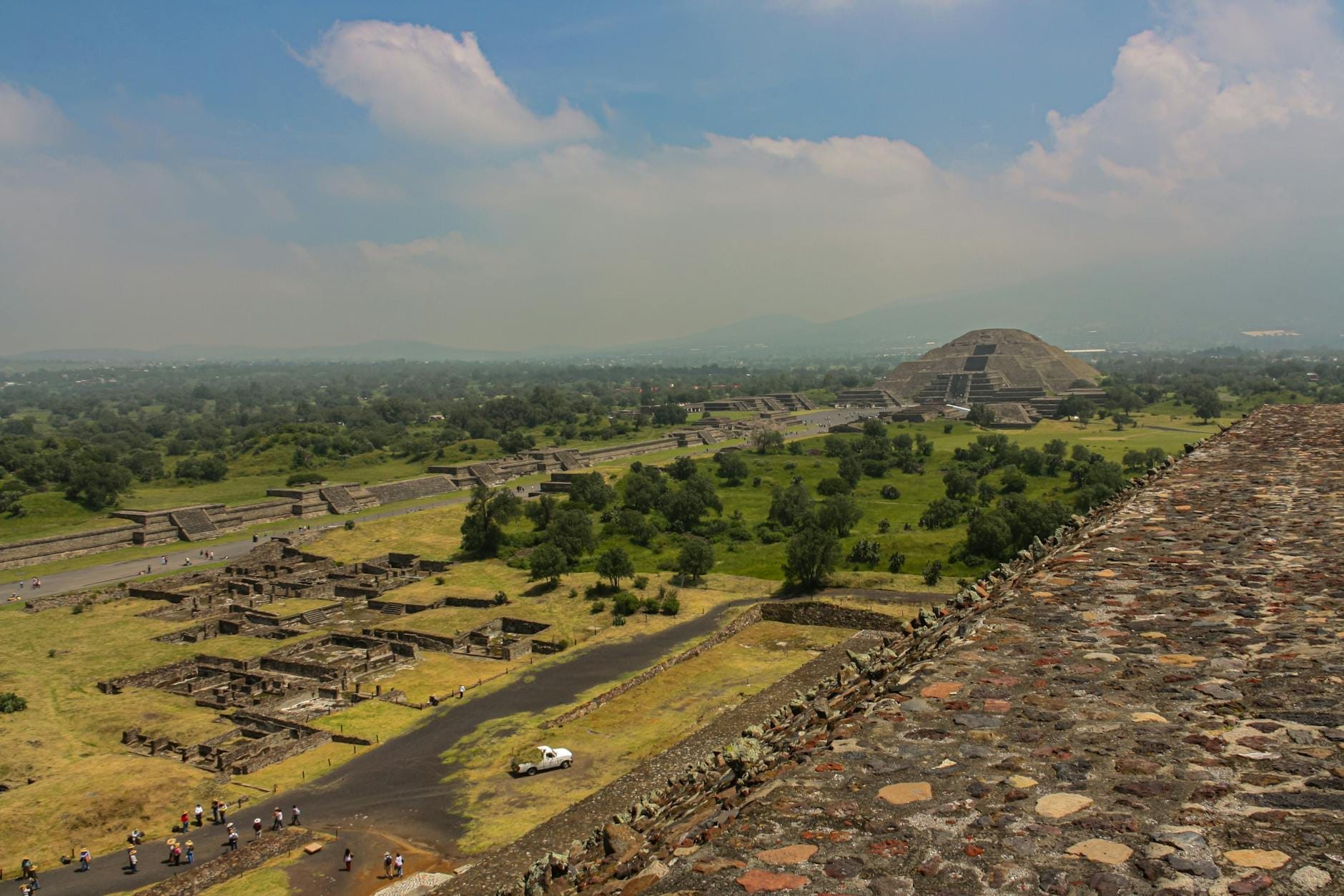 Vista aérea ampla de Teotihuacán com a Pirâmide do Sol ao fundo, vegetação verde ao redor, ruínas e um céu dramático com nuvens.