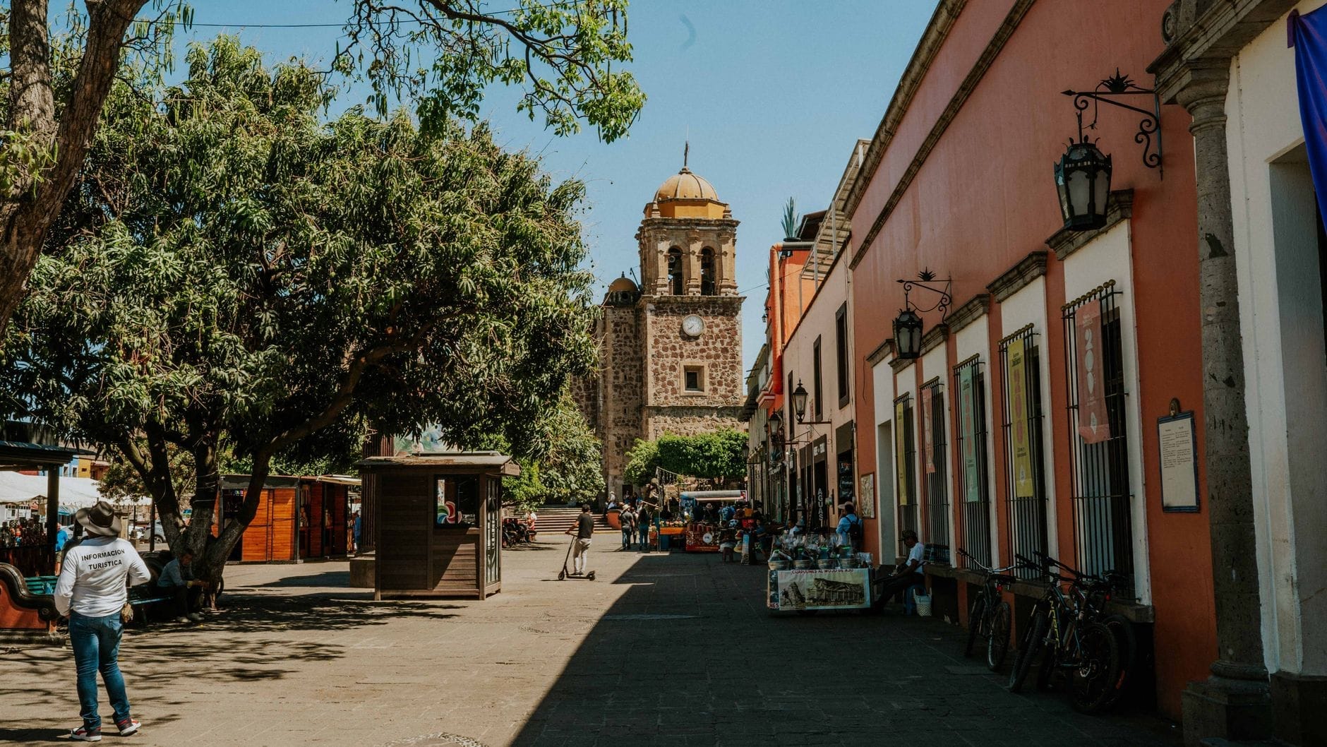 Rua de paralelepípedos em Tequila, México, com coloridos edifícios coloniais, pessoas caminhando, barracas de mercado, árvores e a torre da igreja da cidade ao fundo sob um céu ensolarado.