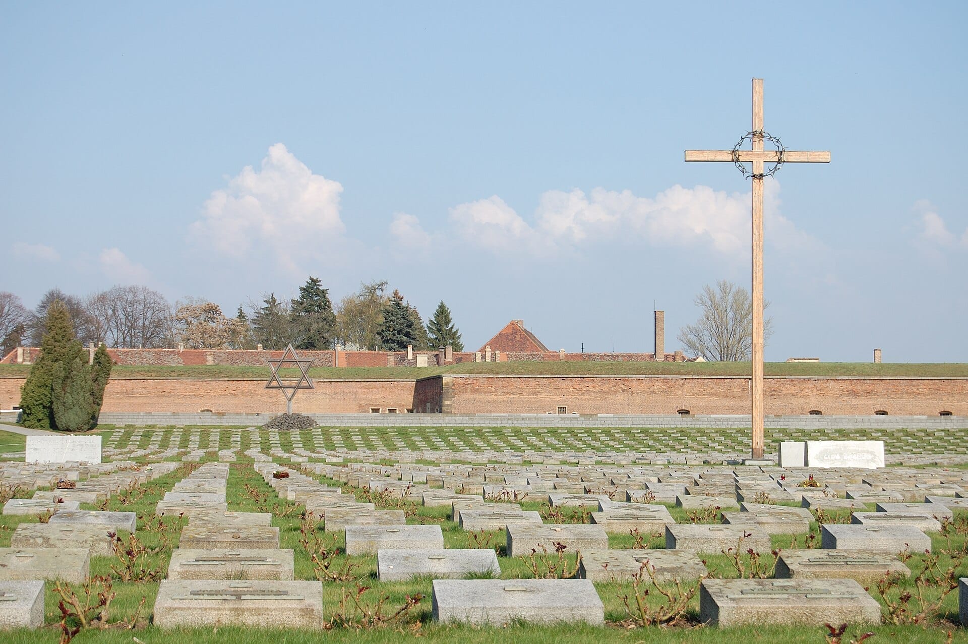 File di lapidi nel cimitero del Memoriale di Terezín con una grande croce di legno e una Stella di David, sotto un cielo azzurro limpido.