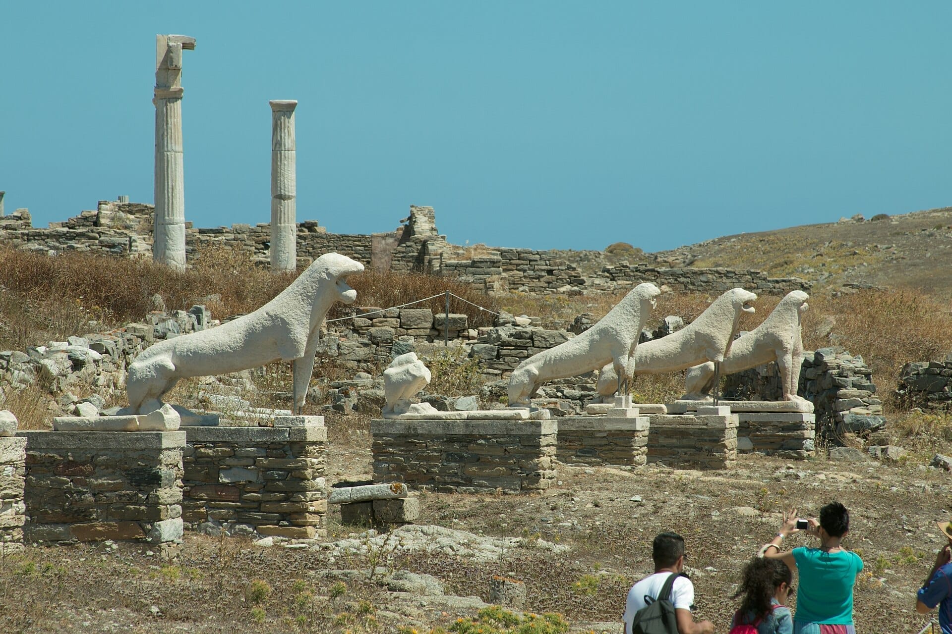Fila de estatuas de leones de mármol en la Terraza de los Leones, Delos, con ruinas antiguas y visitantes bajo un cielo azul brillante.