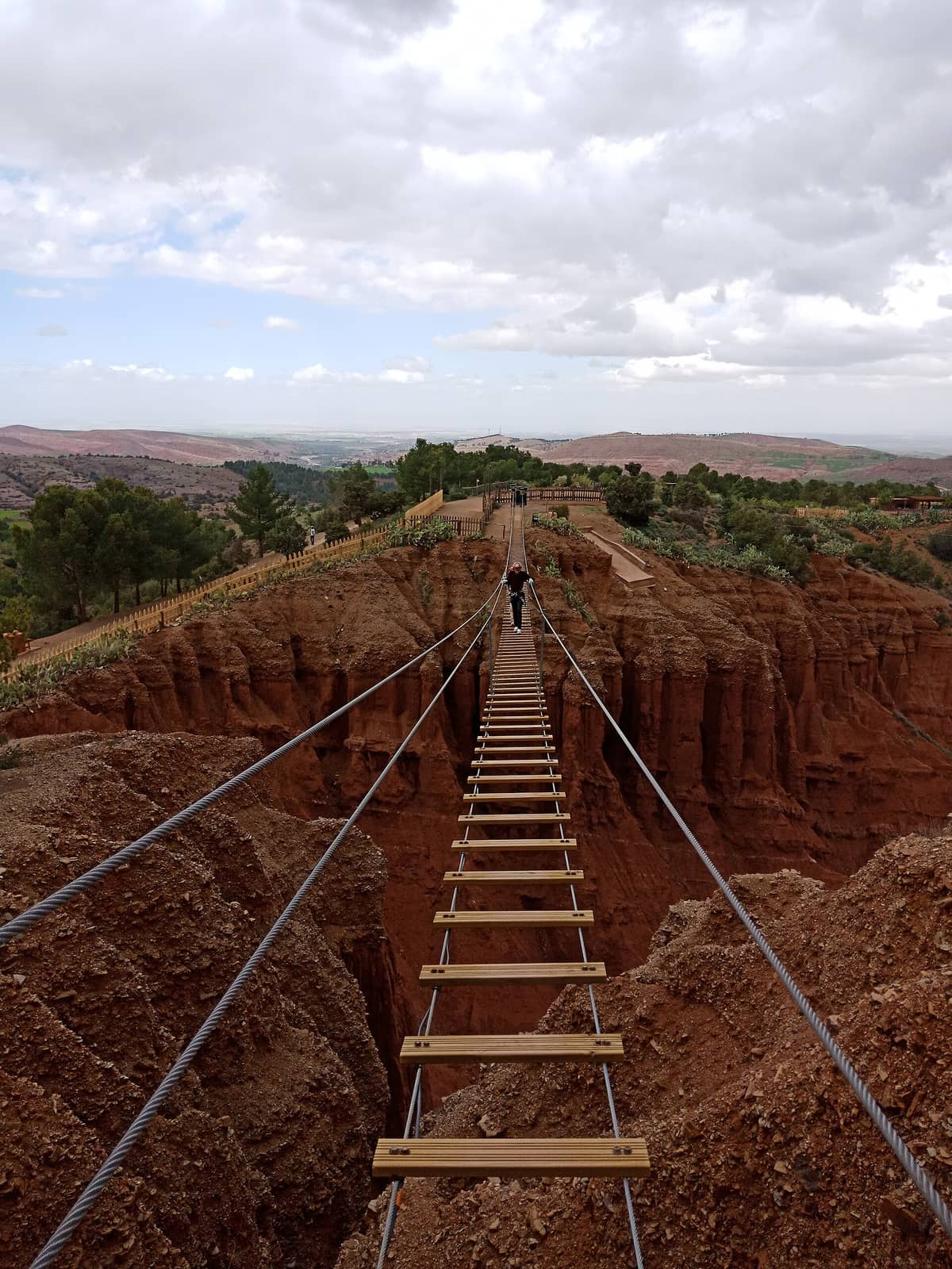 สะพานแขวนไม้ที่ทอดข้ามหุบเขาสีแดงอันน่าตื่นตาใน Terres d'Amanar Adventure Park ล้อมรอบด้วยต้นไม้และภูเขาที่อยู่ไกลออกไปใต้ท้องฟ้าที่มีเมฆปกคลุม