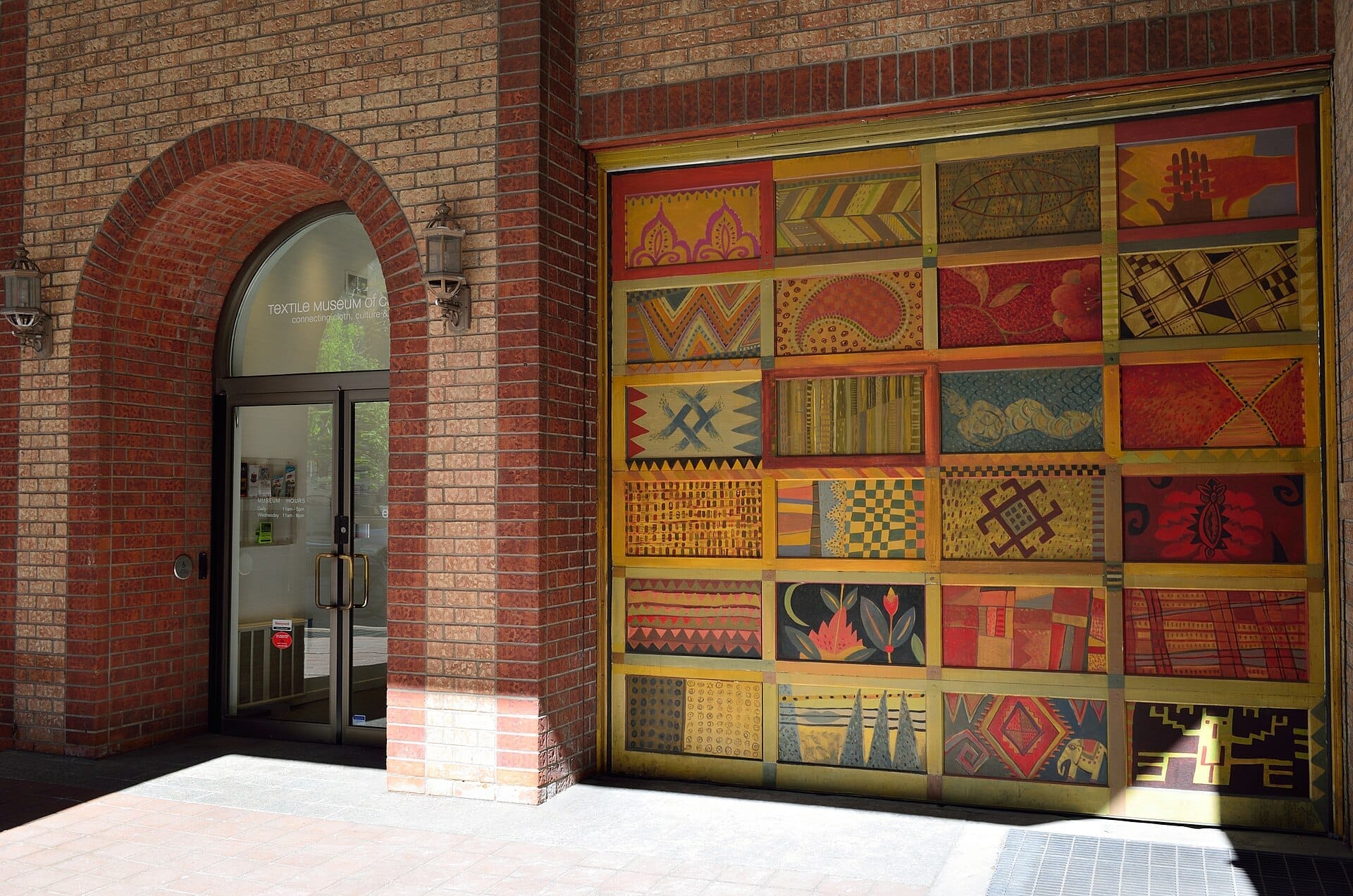 ทางเข้า Textile Museum of Canada ที่มีภาพจิตรกรรมฝาผนังลวดลายสีสันสดใสขนาดใหญ่และประตูกระจกในซุ้มโค้งอิฐ