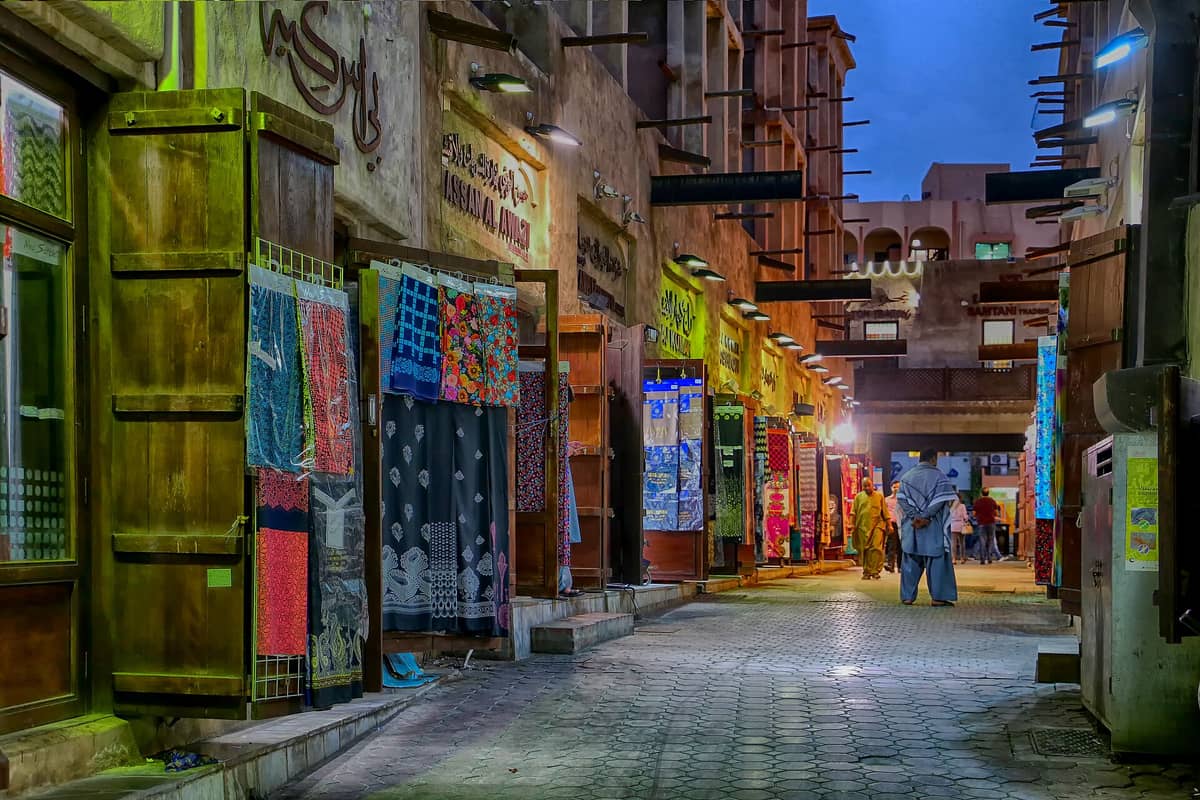 Abendliche Ansicht des Textilsouks in Dubai: Stoffläden säumen beide Seiten einer belebten überdachten Gasse, bunte Textilien sind ausgestellt, und Menschen schlendern hindurch.