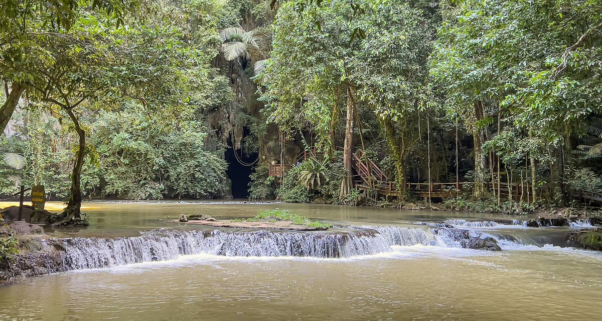 Smaragdgrüner Bach mit kleinen Wasserfällen, der durch dichten Dschungel fließt, mit Holzstegen und Höhleneingang im Than Bok Khorani Nationalpark, Krabi.