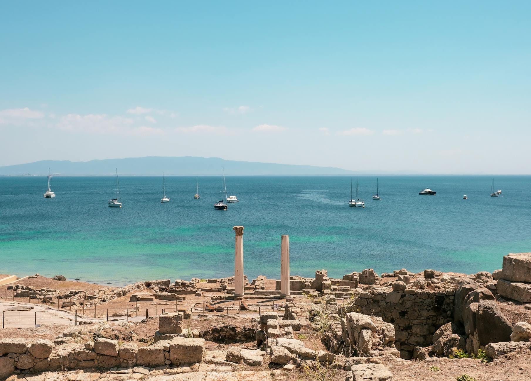 Antike Steinruinen mit zwei stehenden römischen Säulen mit Blick auf türkisblaues Meer und Segelboote, mit einer fernen Küstenlinie unter strahlendem blauen Himmel.