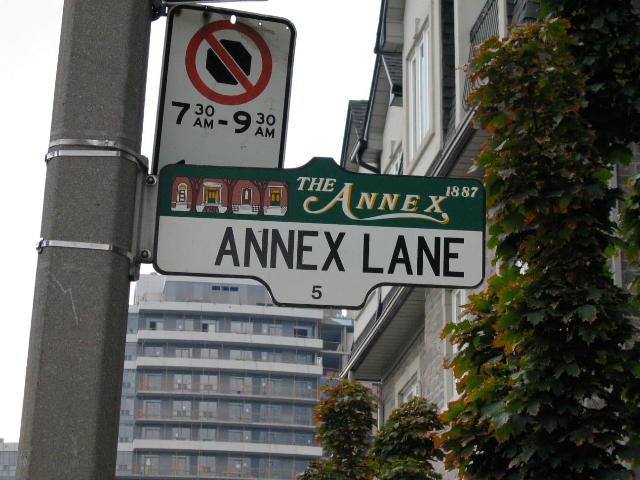 ภาพระยะใกล้ของป้ายถนน Annex Lane ในย่าน The Annex เมือง Toronto โดยมีอาคารอพาร์ตเมนต์ ต้นไม้สีเขียว และอาคารสมัยใหม่เป็นฉากหลัง