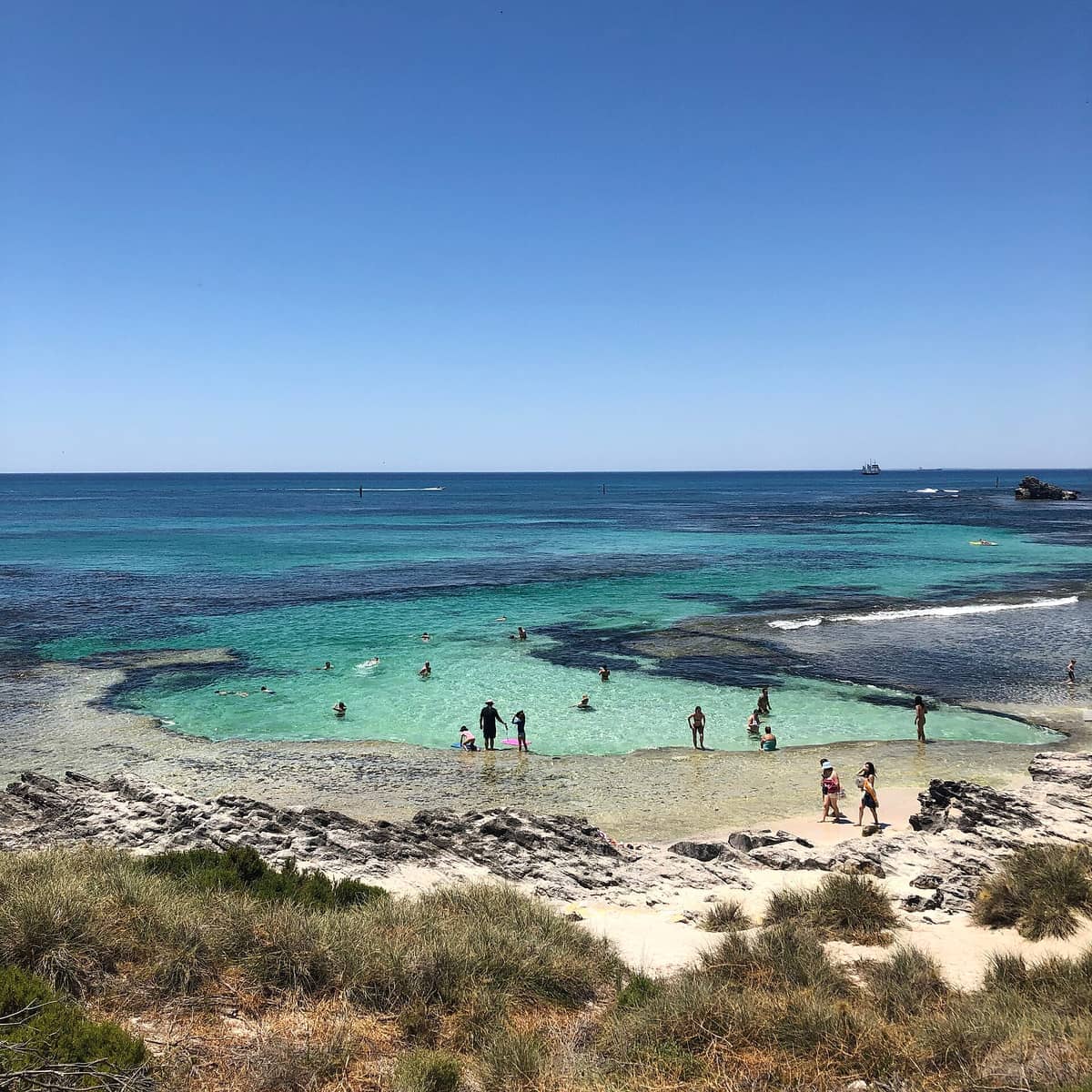 Besucher schwimmen und entspannen sich im klaren, rifgeschützten Wasser von The Basin auf Rottnest Island, umgeben von Felsvorsprüngen und Sandstrand.