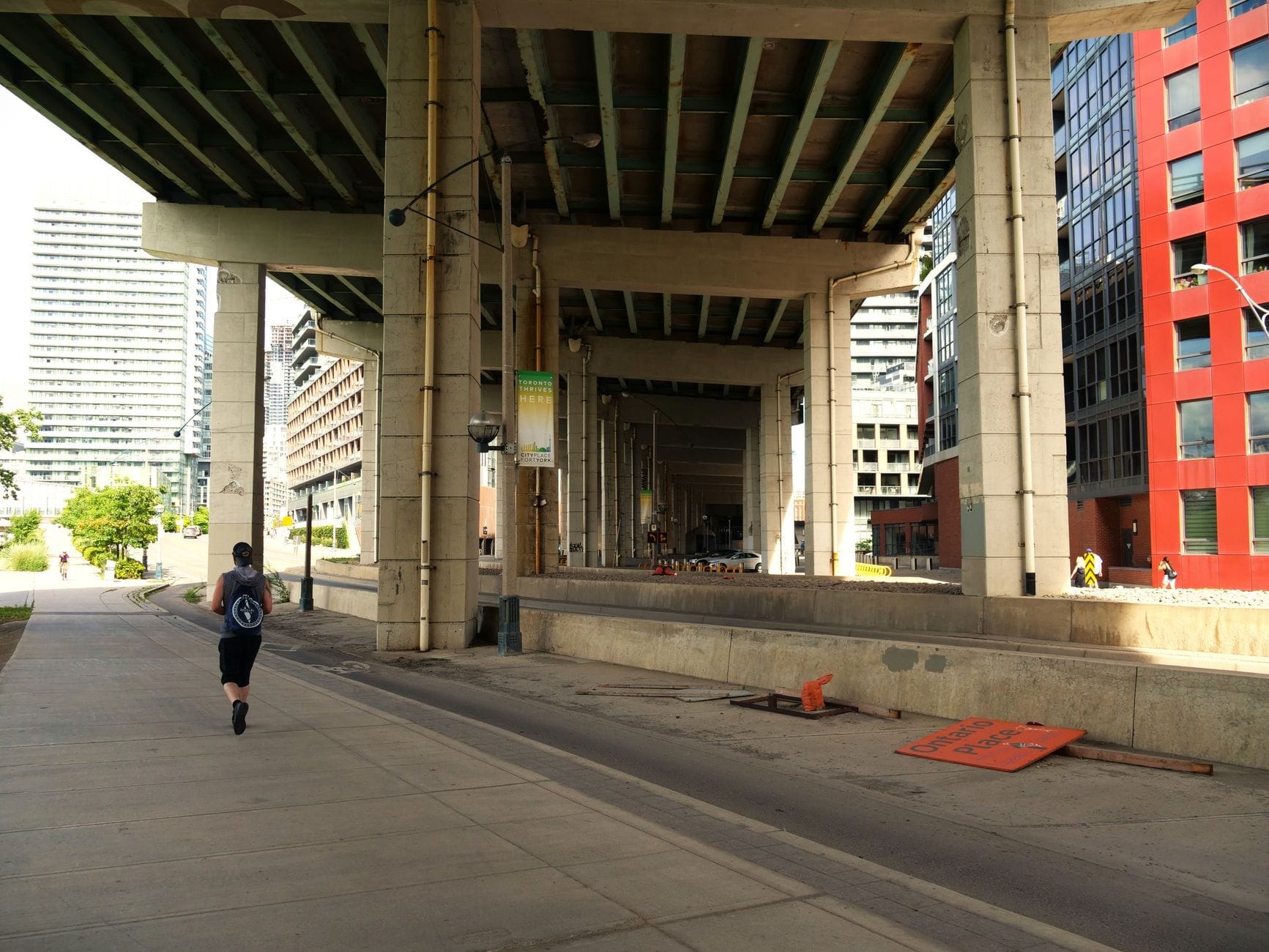 มุมกว้างใต้สะพาน Gardiner Expressway ในโตรอนโต มีทางเดินคนเมืองและจักรยาน พร้อมนักวิ่งและอาคารสมัยใหม่ทั้งสองฝั่ง