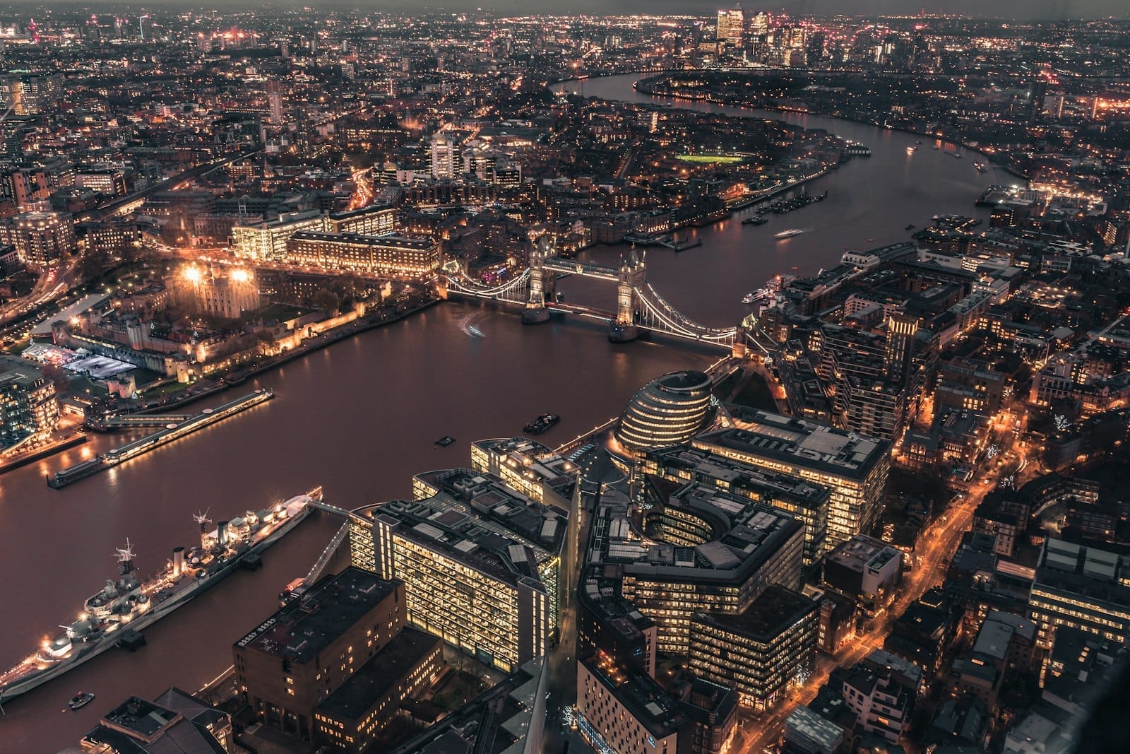 ภาพมุมสูงยามค่ำของ The City of London ตึกสำนักงานเปิดไฟระยิบระยับ เห็น Tower Bridge และโค้งแม่น้ำเทมส์ข้ามสถาปัตยกรรมเก่าใหม่ในสแควร์ไมล์