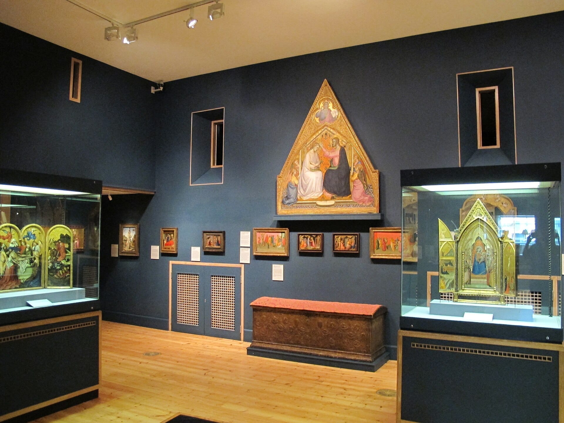 Sala della galleria presso The Courtauld Gallery con pale d'altare medievali illuminate, dipinti religiosi e pavimenti in legno ornati sotto una soffusa illuminazione espositiva.