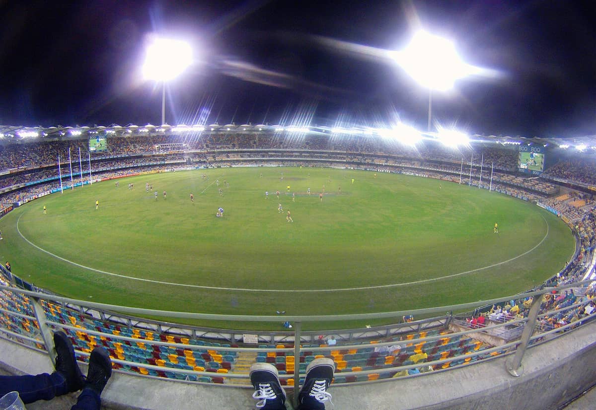 Vista grandangolare dell'interno del Gabba di notte durante una partita di AFL, con il campo brillantemente illuminato, gli spalti pieni di tifosi e i pali della porta ben visibili.