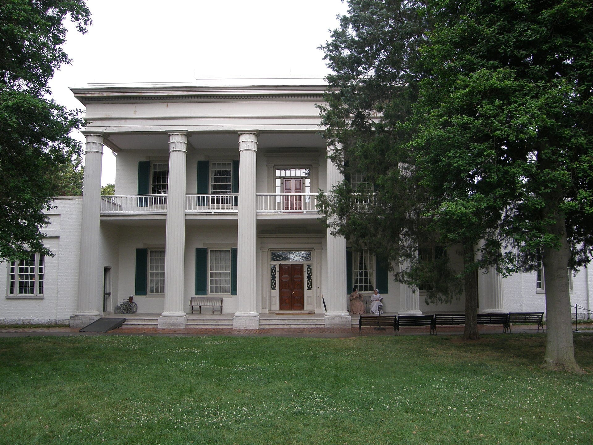 Vista frontal de la mansión Hermitage de Andrew Jackson, con grandes columnas blancas, contraventanas verdes y rodeada de frondosos árboles y césped en un día nublado.