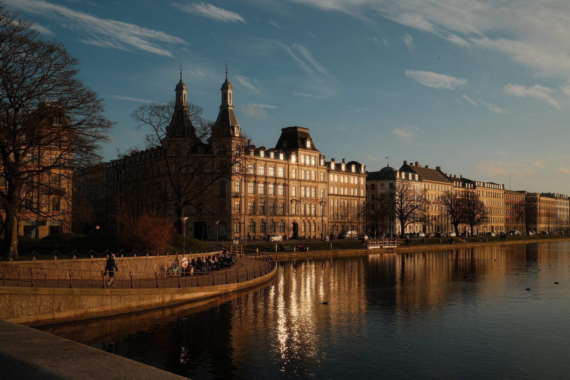 Vista del tardo pomeriggio sul lungolago di Copenaghen: persone in passeggiata lungo la riva, eleganti edifici storici riflessi nell'acqua calma sotto un cielo dorato.