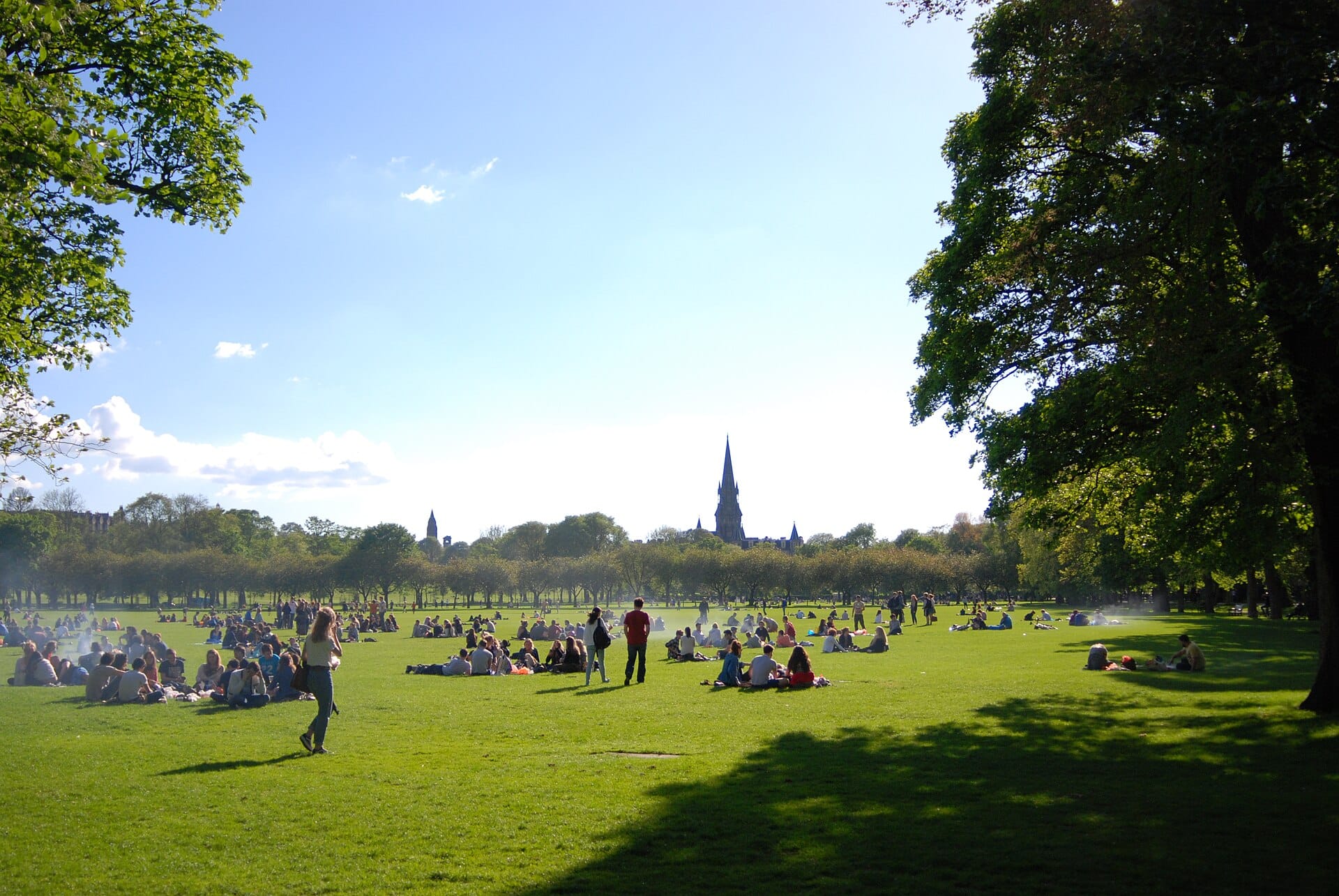 Ein sonniger Tag im Meadows-Park in Edinburgh: Menschen entspannen auf dem Gras, im Hintergrund Laubbäume und die Türme einer Kirche.