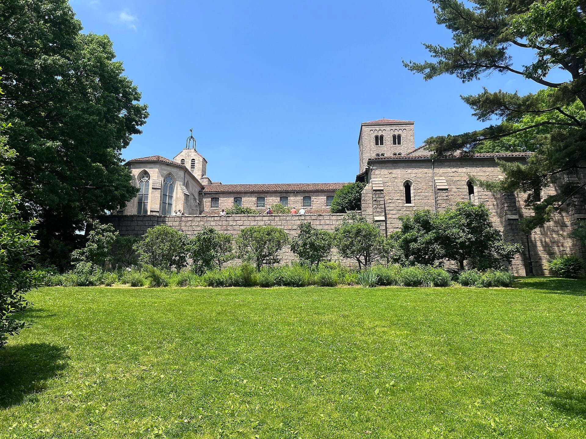 Vista esterna di The Met Cloisters con mura in pietra medievali, torri, giardini paesaggistici e alberi frondosi sotto un cielo azzurro brillante.
