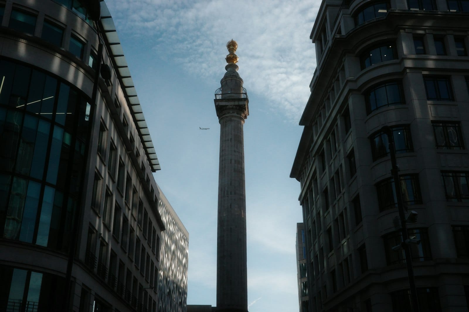 Il Monument to the Great Fire of London si erge tra due moderni edifici per uffici sotto un cielo azzurro brillante con nuvole sparse.