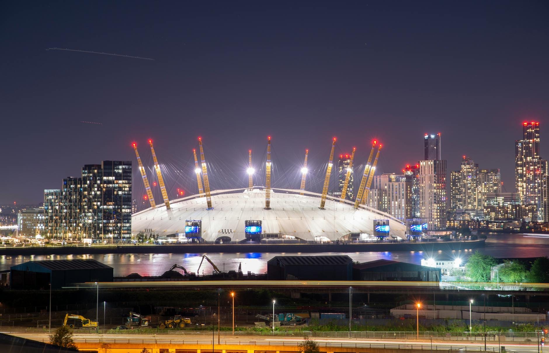 La O2 Arena illuminata di notte con i grattacieli di Londra sullo sfondo, vista dal fiume sotto un cielo sereno.
