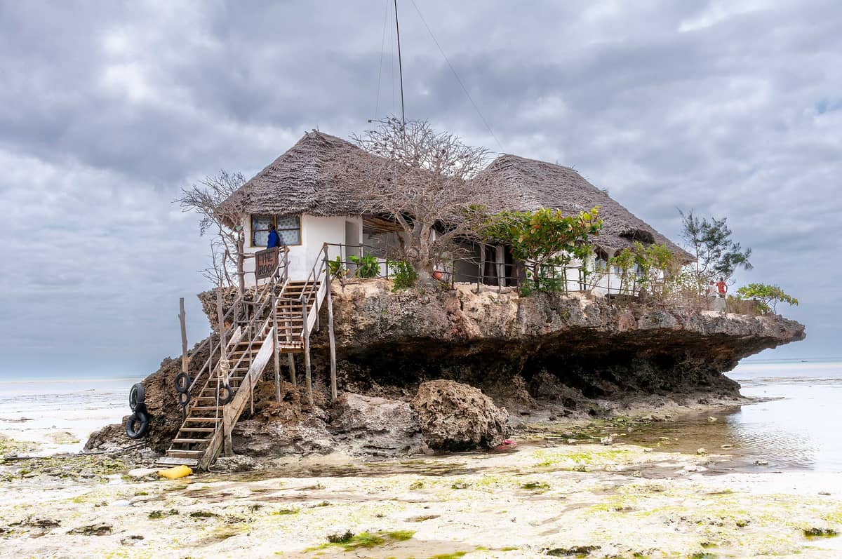 The Rock Restaurant Zanzibar na małej koralowej skale z drewnianymi schodkami, otoczony płytką wodą oceaniczną podczas odpływu i zachmurzonym niebem powyżej.
