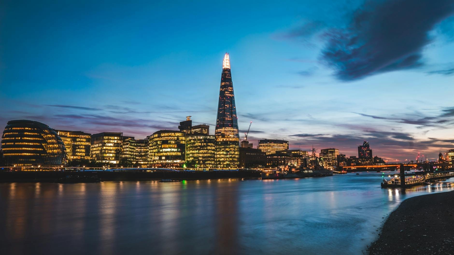 Il grattacielo The Shard illuminato al crepuscolo, che svetta sullo skyline di Londra con i riflessi delle luci sul Tamigi, sotto un cielo vibrante di blu e viola.